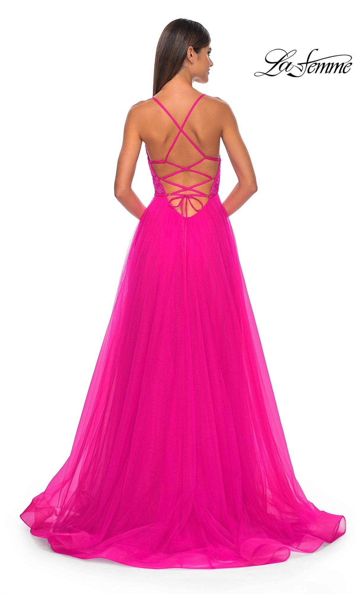 A woman models the La Femme Deep V-Neck Long Tulle Prom Gown 32059, featuring a lace bodice, strappy open back, and flowing pink skirt. She stands against a white backdrop with the